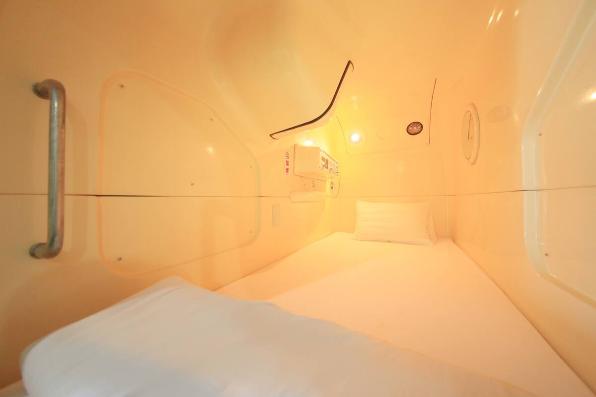 A small, compact room with a single bed, a bedside table, and a wall-mounted control panel.