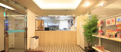 Un hall de hotel con mostrador de recepción, ascensor de vidrio, planta y bar de aperitivos.