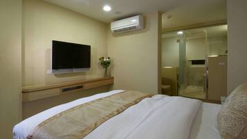 Hotelkamer met een bed, een flatscreen televisie aan de muur, een airconditioning, een kleintje tafeltje met een bloemenvaas, en een glazen schuifdeur naar een douchegedeelte.