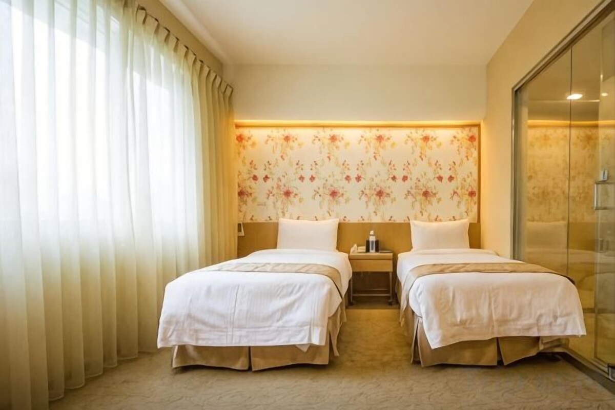A hotel room with two beds, a nightstand, a lamp, and a large window with curtains.