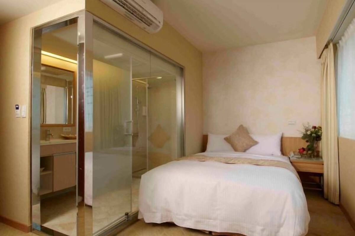 A hotel room with a bed, a glass door leading to a bathroom, and a nightstand with a lamp.