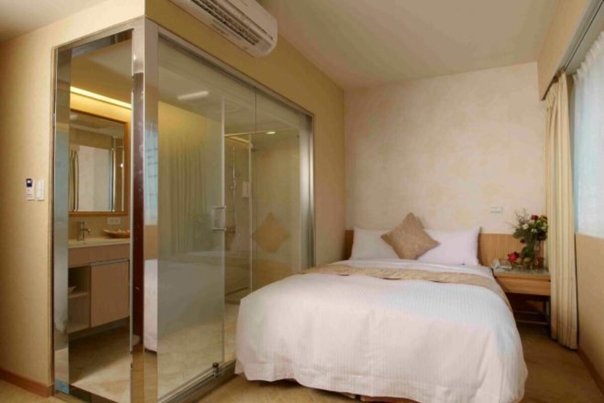 A hotel room with a bed, a glass shower enclosure, a bedside table with a lamp, and a window with curtains.