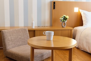 Ett hotellrum med ett nattduksbord i trä, en vit mugg, en liten vas med blommor, en säng med vita sängkläder och en randig vägg.