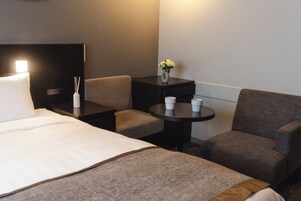Habitación de hotel con cama, dos sillones, una mesita con tazas, un jarrón con flores y un televisor de pared.