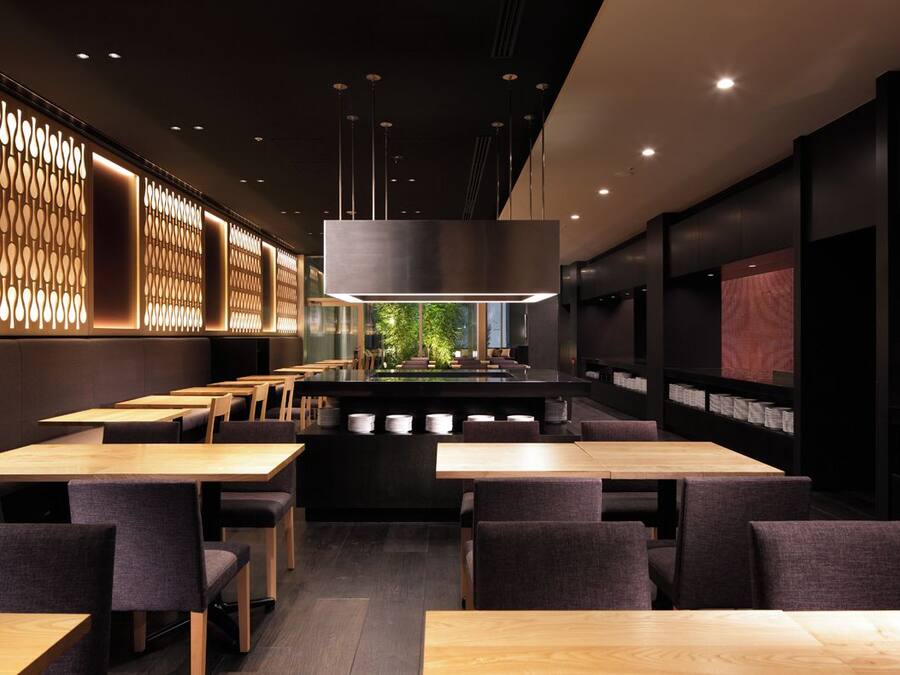 Un intérieur de restaurant moderne, avec plusieurs tables et chaises, un luminaire central et une vue sur la verdure à l’extérieur.