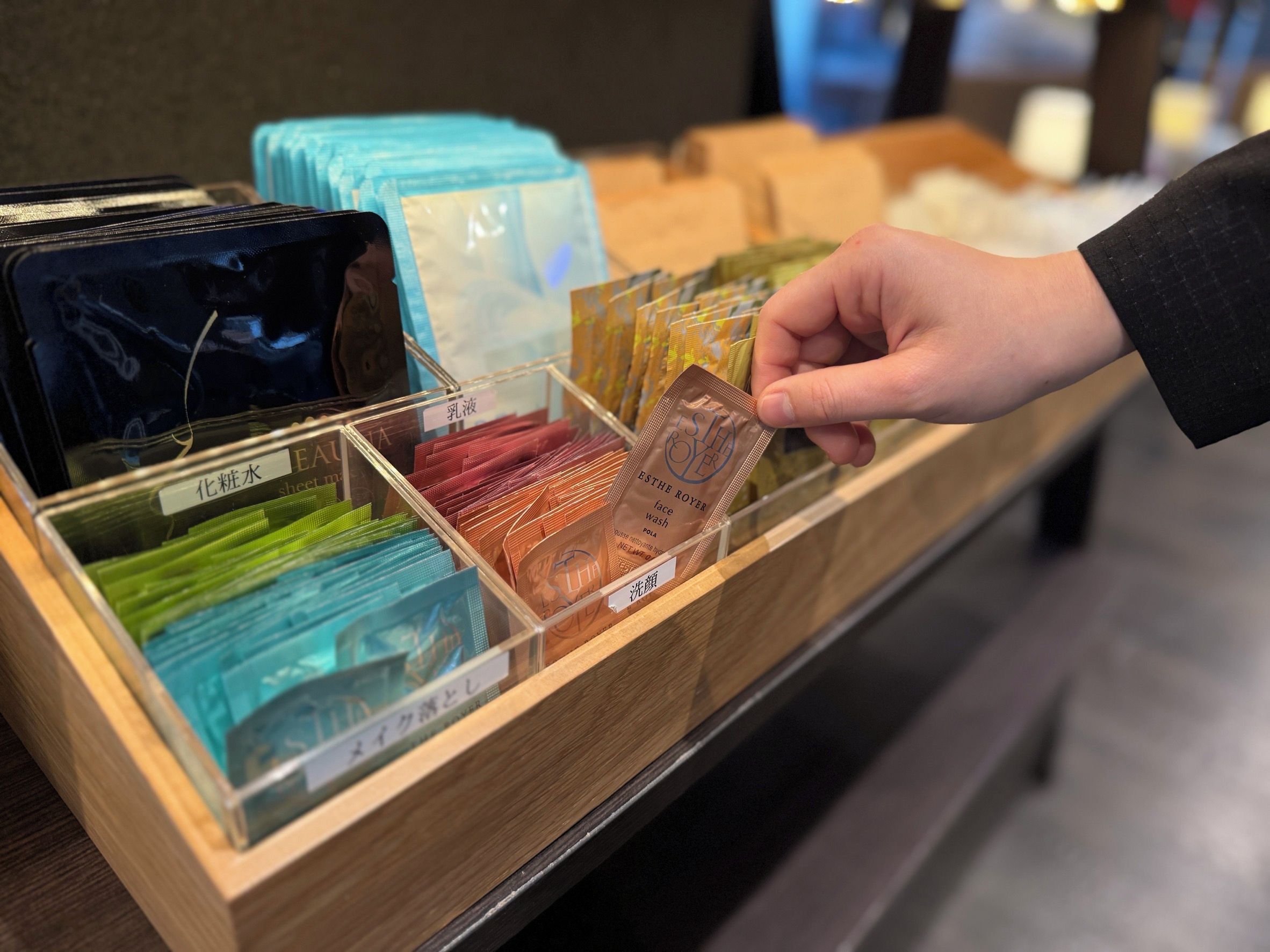 A hand selecting a face wash product from a wooden display case containing various skincare items.