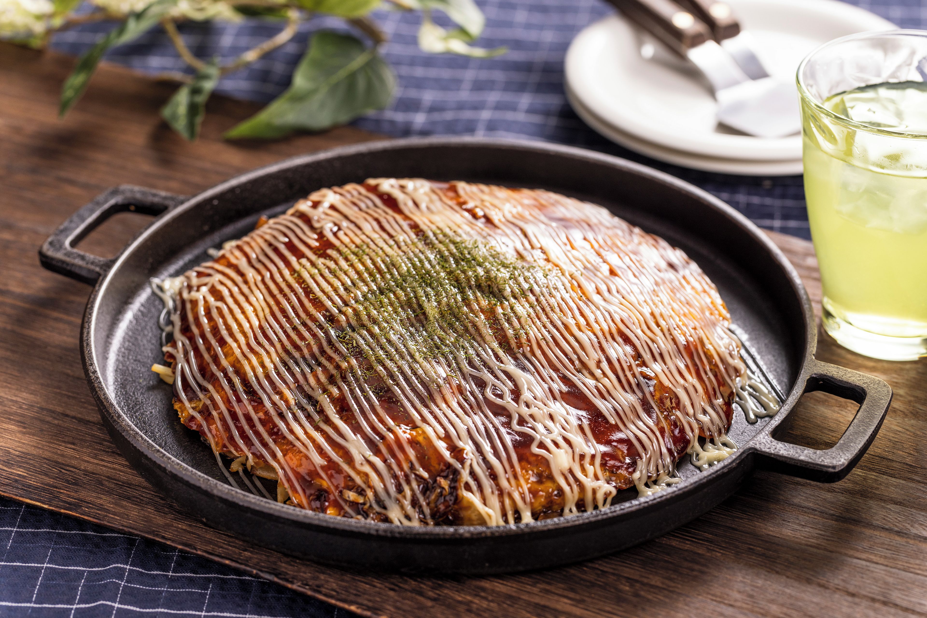 Une poêle en fonte contenant un plat nappé de sauce et recouvert de fromage, posée sur une table en bois recouverte d’une nappe à carreaux.