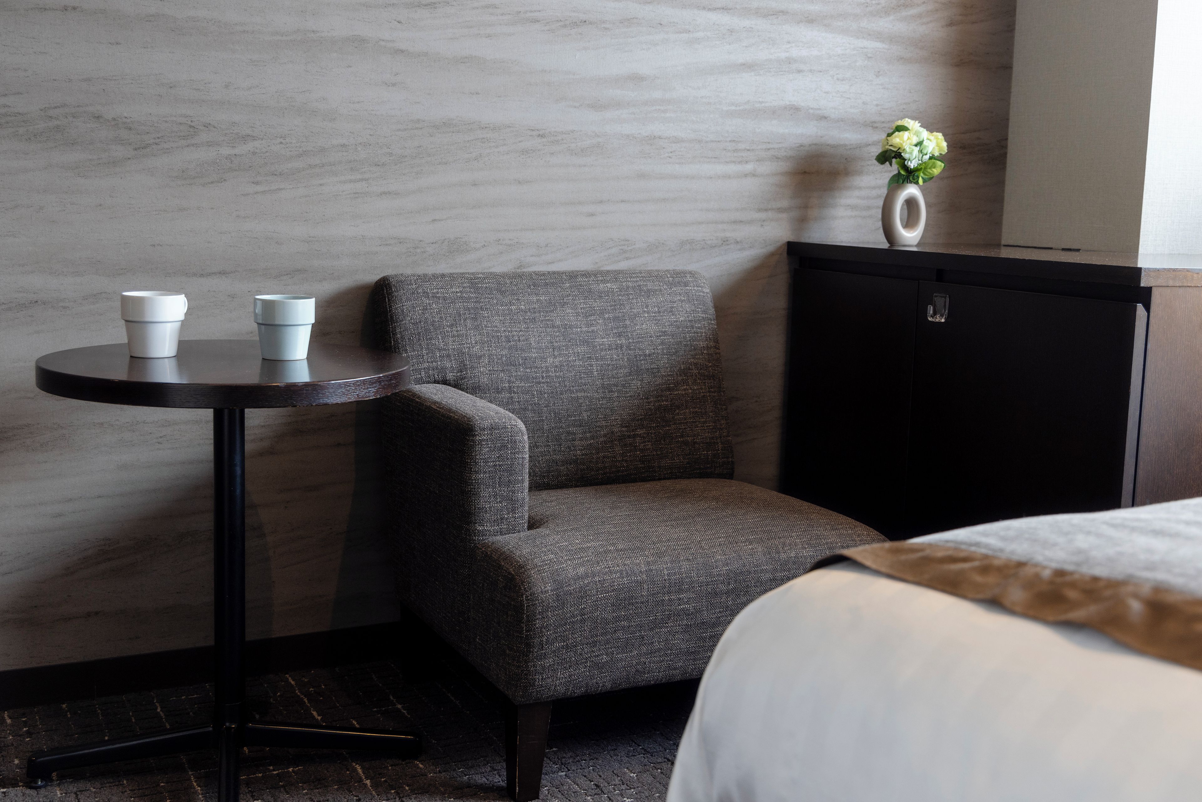 A hotel room with a grey upholstered armchair, a small round table with two white cups, a black cabinet, and a vase with flowers on top.