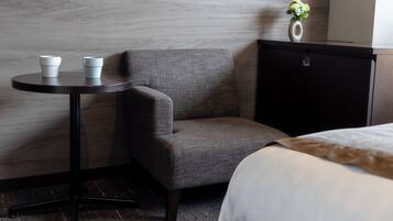 A hotel room with a grey upholstered armchair, a small round table with two white cups, a black cabinet, and a vase with flowers on top.