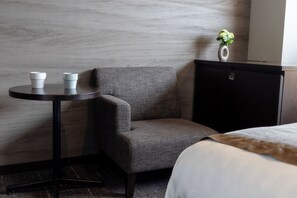 A hotel room with a grey upholstered armchair, a small round table with two white cups, a black cabinet, and a vase with flowers on top. - Hiroshima Washington Hotel (Hiroshima)
