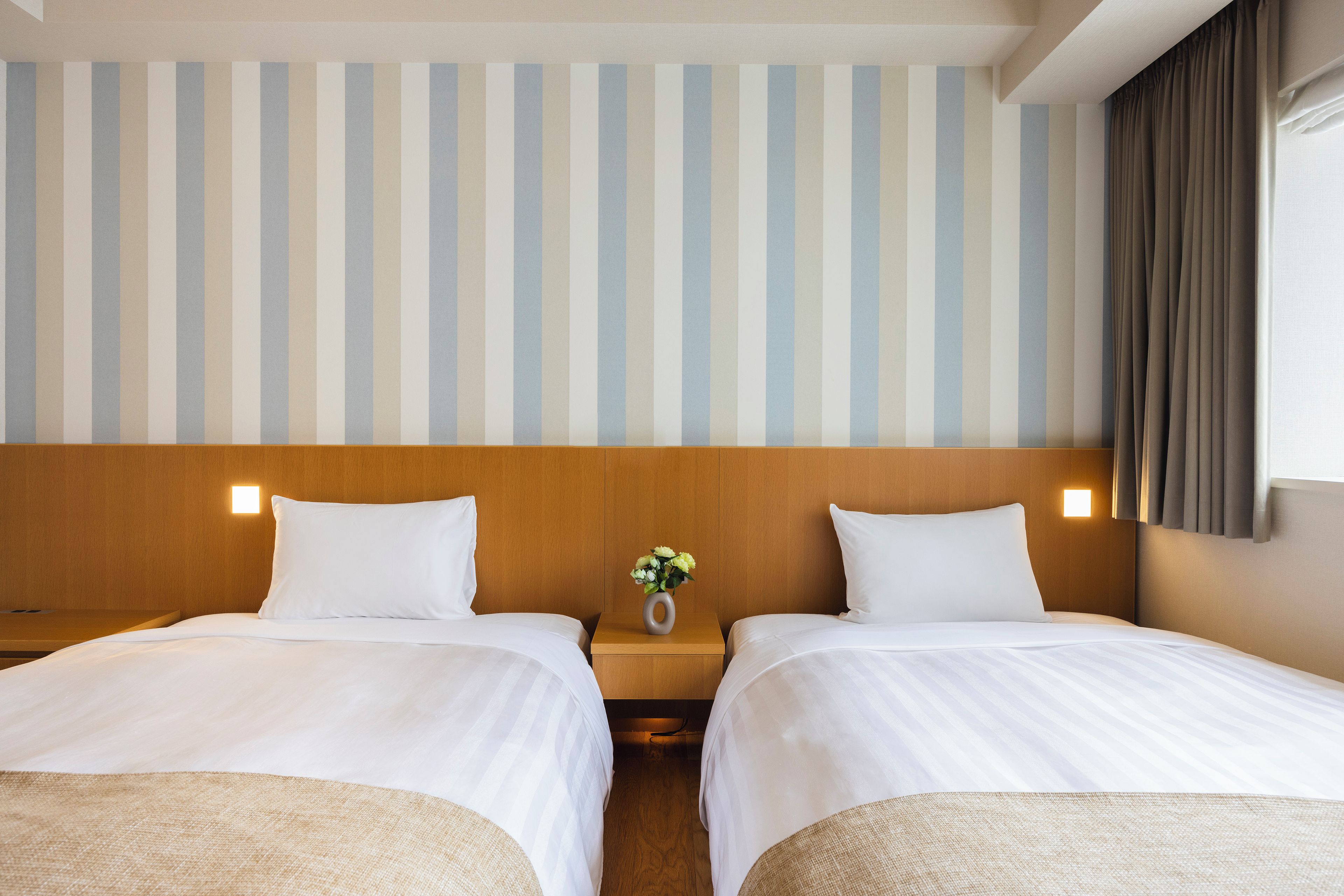 A hotel room with two beds, a wooden headboard, a striped wall, and a small table with a vase of flowers.