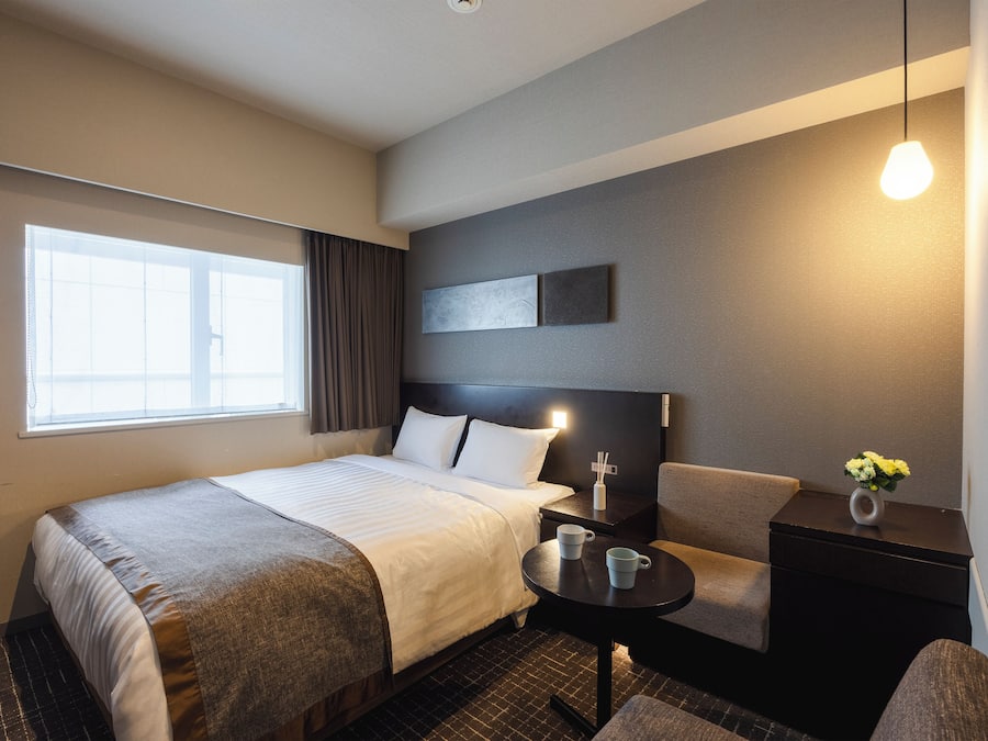 A hotel room with a bed, a small table, a chair, a nightstand, and a window.