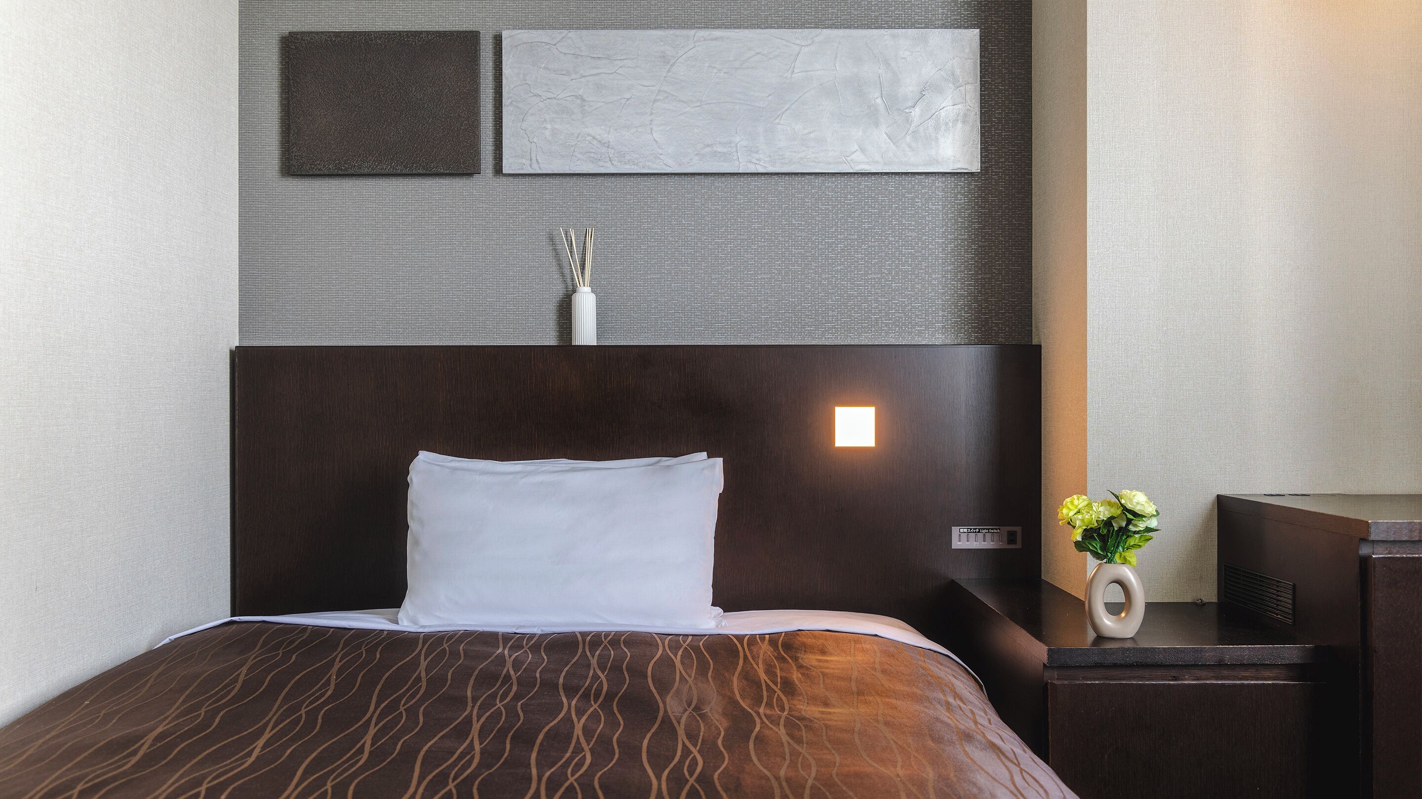 Un lit bien fait, avec une tête de lit sombre, une table de chevet sur laquelle se trouve une lampe, et un luminaire fixé au mur.