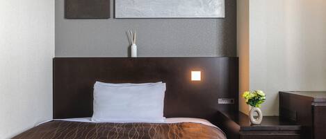 Un lit bien fait, avec une tête de lit sombre, une table de chevet sur laquelle se trouve une lampe, et un luminaire fixé au mur.