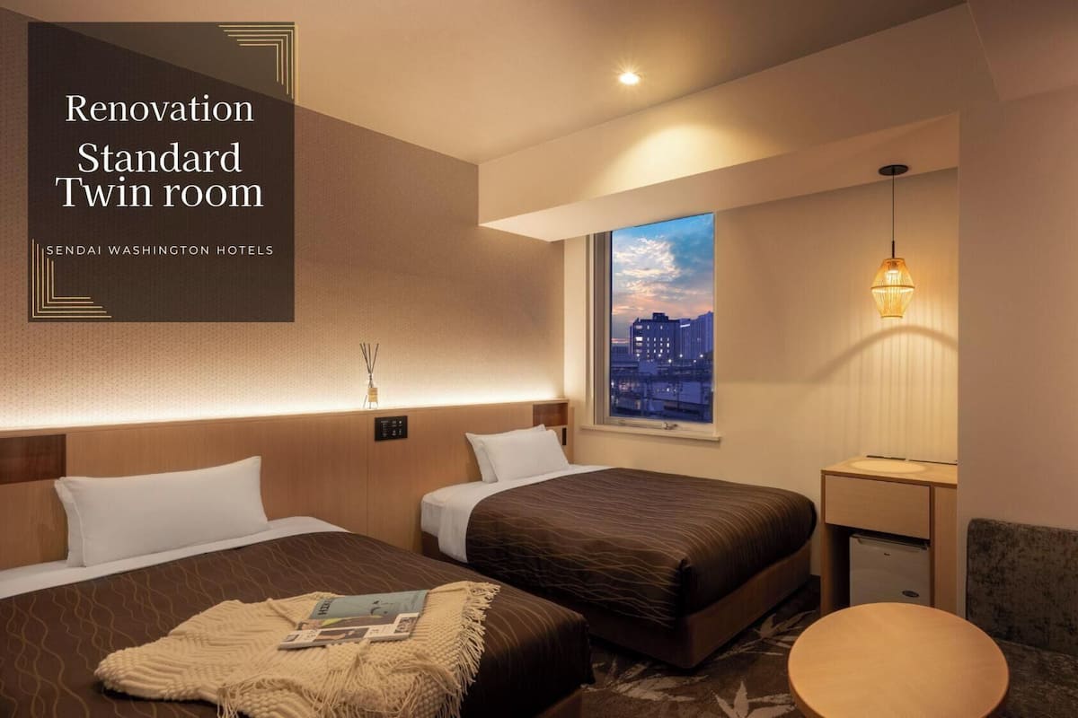 A renovated standard twin room with two beds, a small table, a chair, a nightstand, a lamp, and a city view.