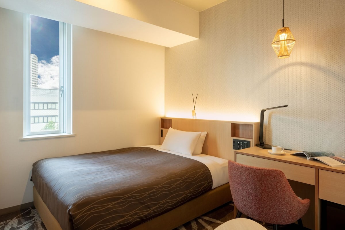 Habitación de hotel con cama, escritorio con silla, lámpara, ventana con vista a edificios, y lámpara de pared.