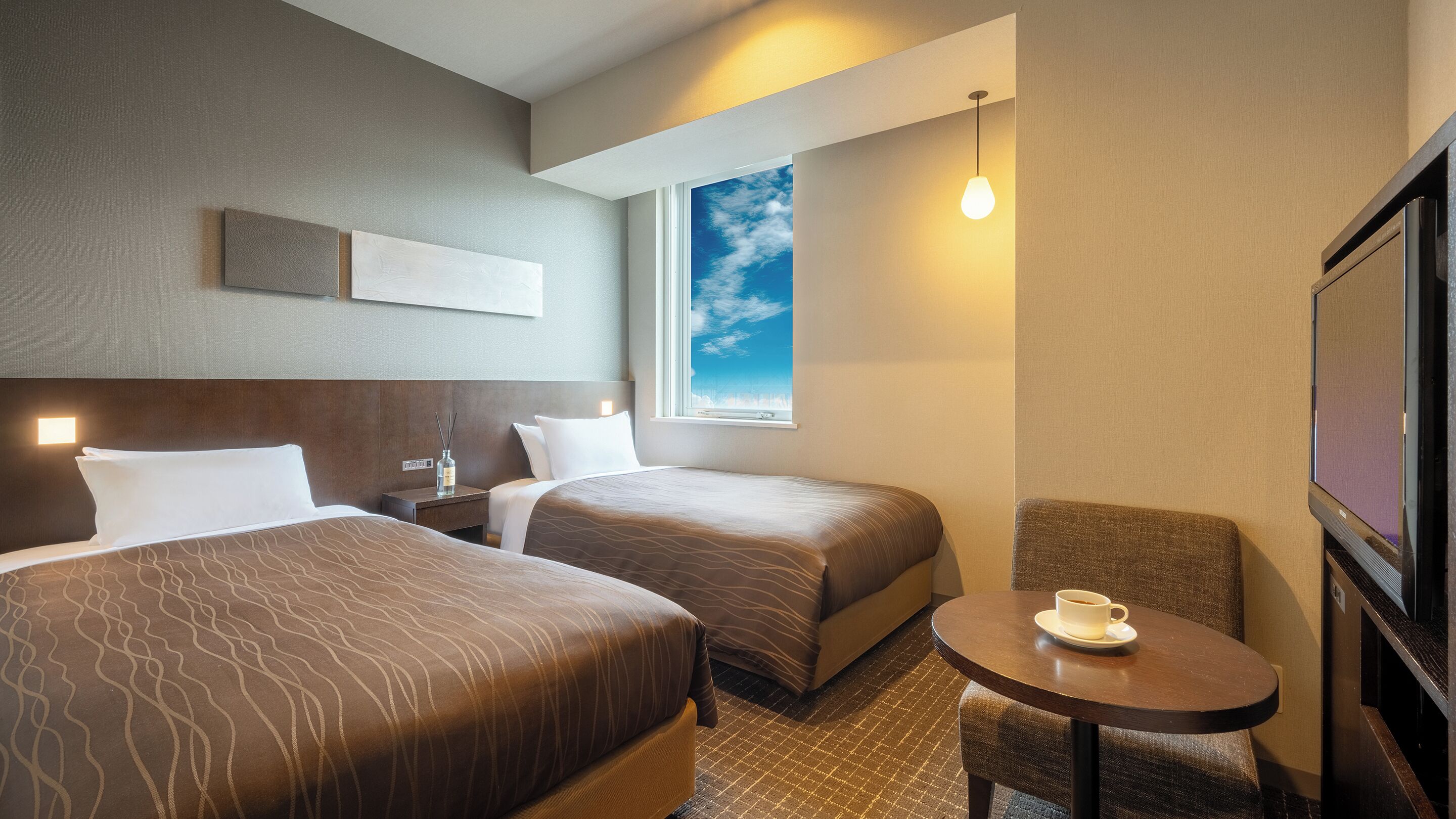 Hotelkamer met twee bedden, een klein tafeltje met een kopje, een raam met uitzicht op de lucht en een flatscreen televisie.