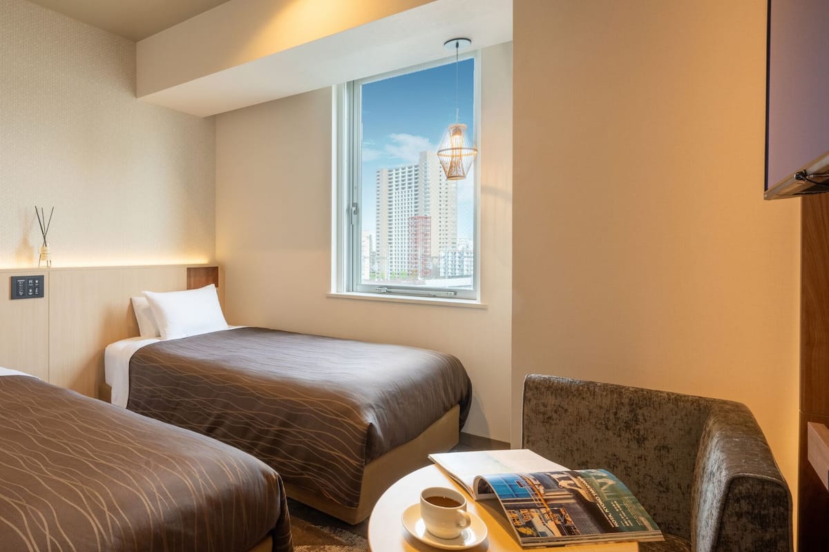 Habitación de hotel con dos camas, una ventana con vista a la ciudad, una lámpara y una mesa con un libro y una taza.