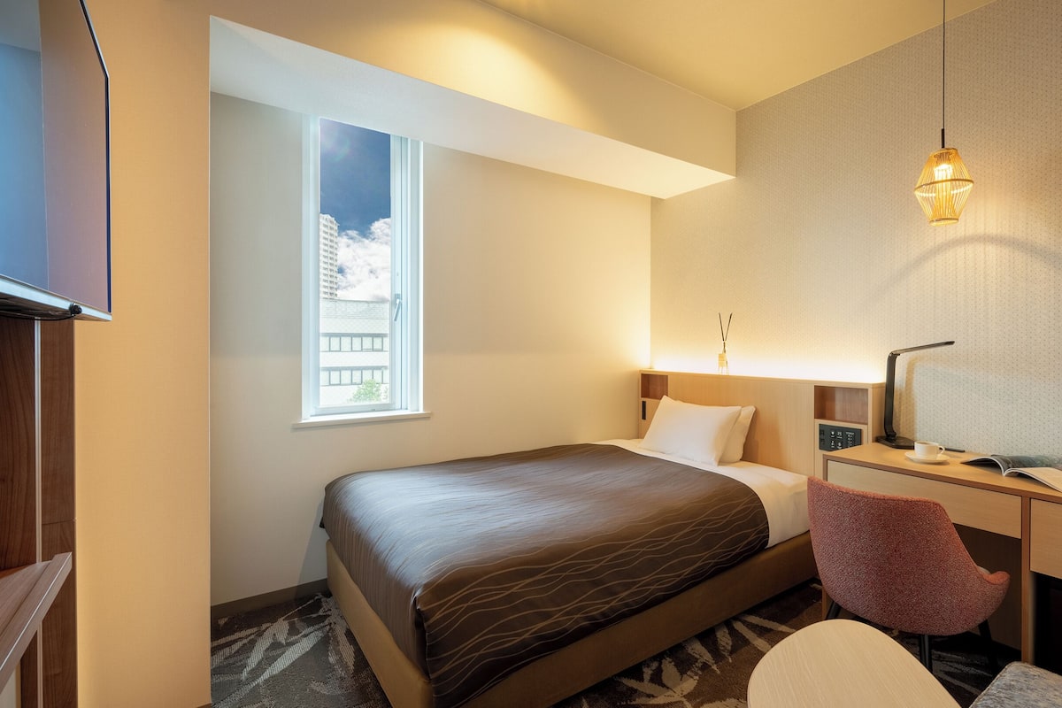 Una habitación de hotel con cama, escritorio, silla, lámpara, ventana y una lámpara fijada a la pared.