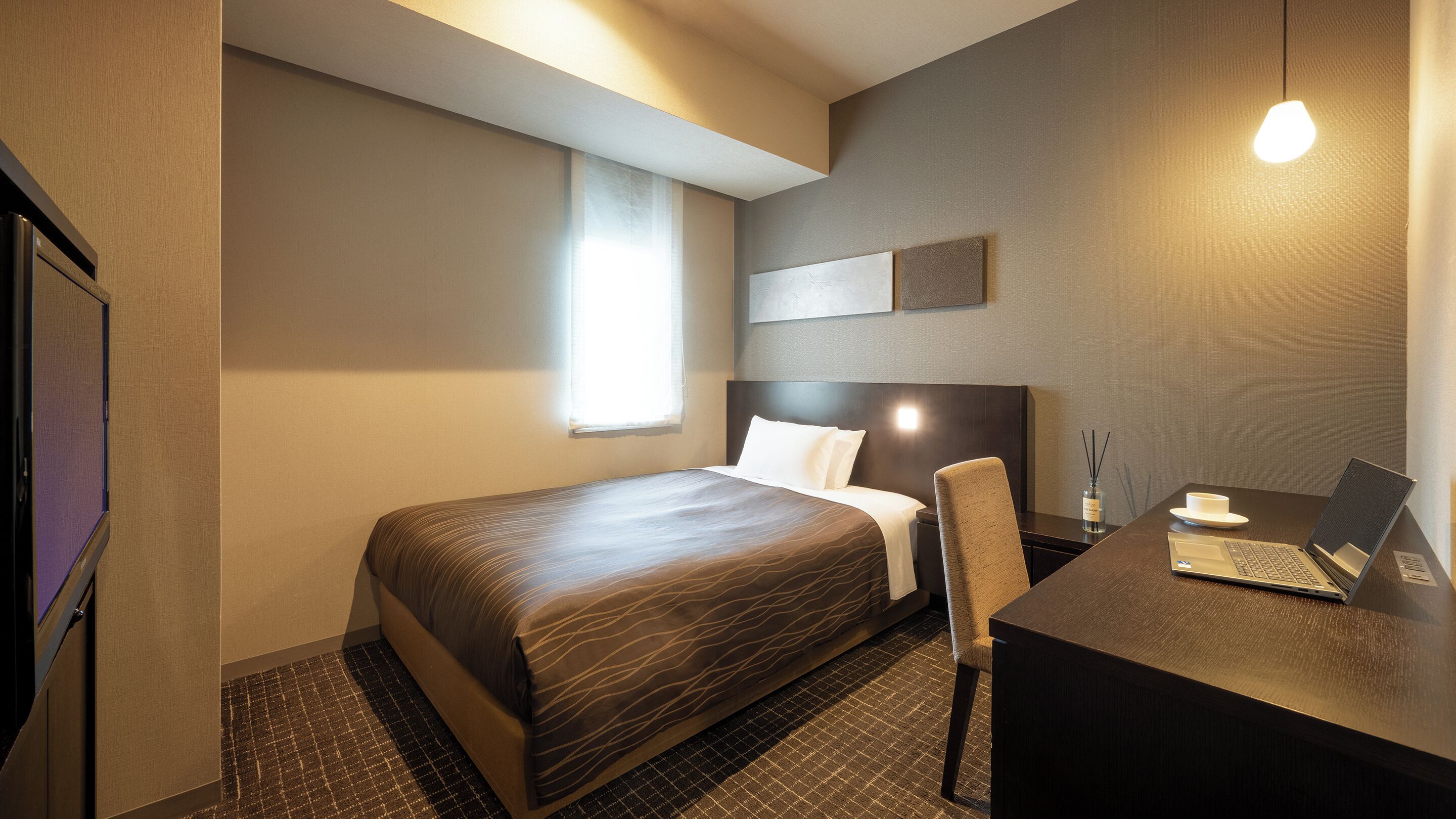 A hotel room with a bed, a desk with a laptop, a chair, a small table with a cup and a bottle, and a window with curtains.