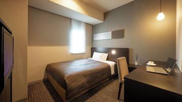 A hotel room with a bed, a desk with a laptop, a chair, a small table with a cup and a bottle, and a window with curtains.