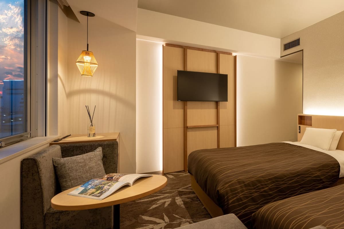 Una habitación de hotel con cama, televisión, escritorio con lámpara y ventana con vista a la ciudad.