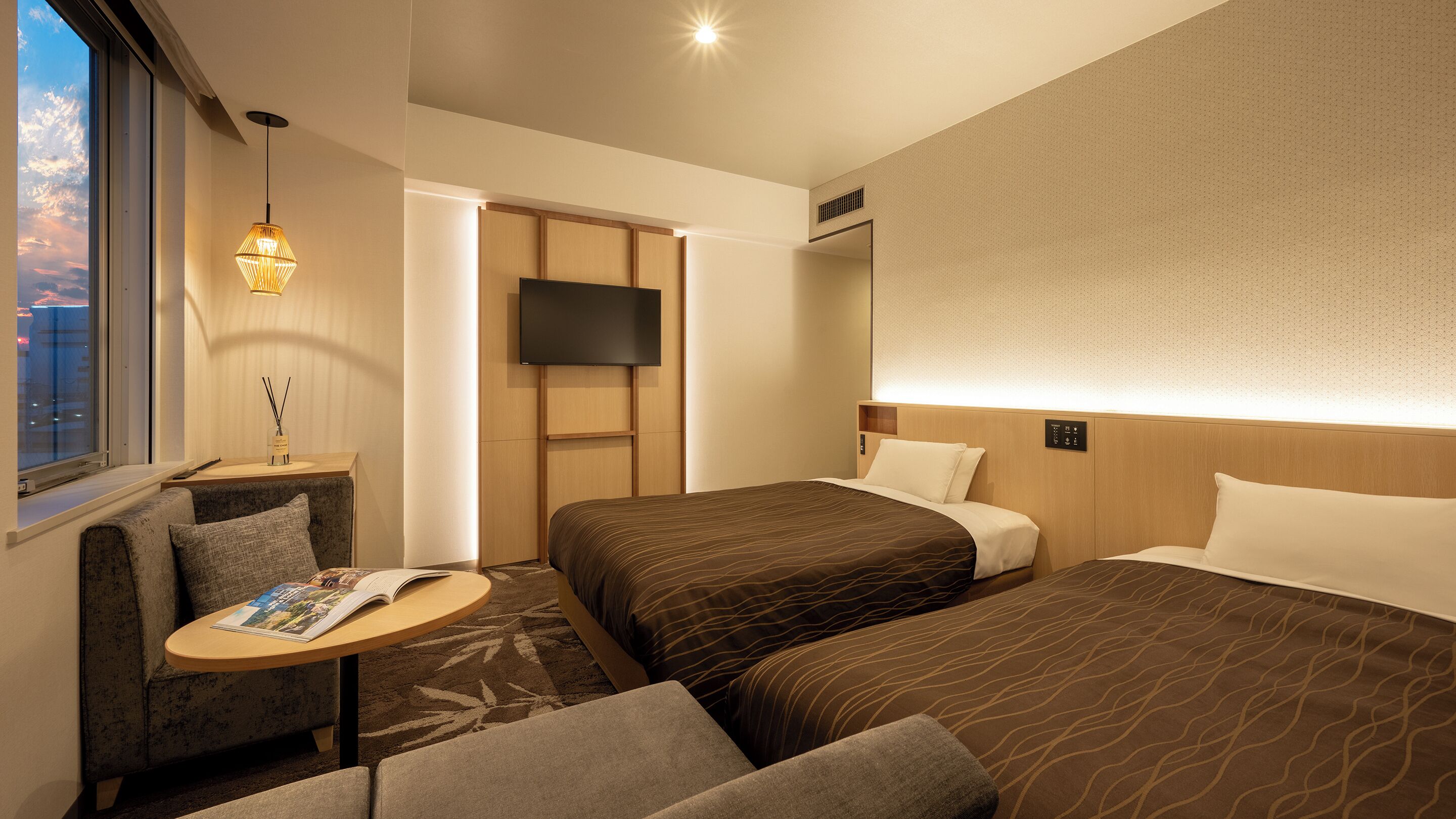 A hotel room with two beds, a TV, a small table with a magazine, and a window with a view of the city.