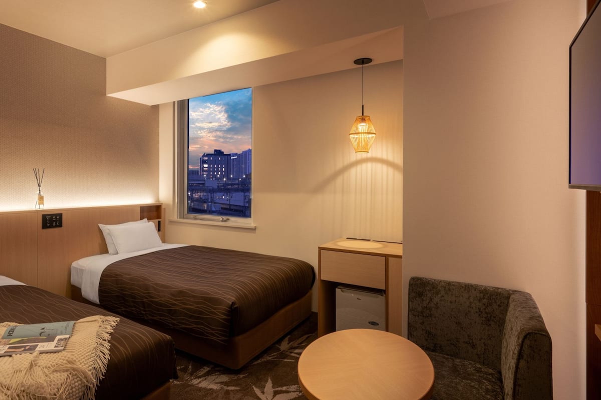 Habitación de hotel con cama, una mesita, una silla, ventana con vista a la ciudad y una lámpara fijada a la pared.