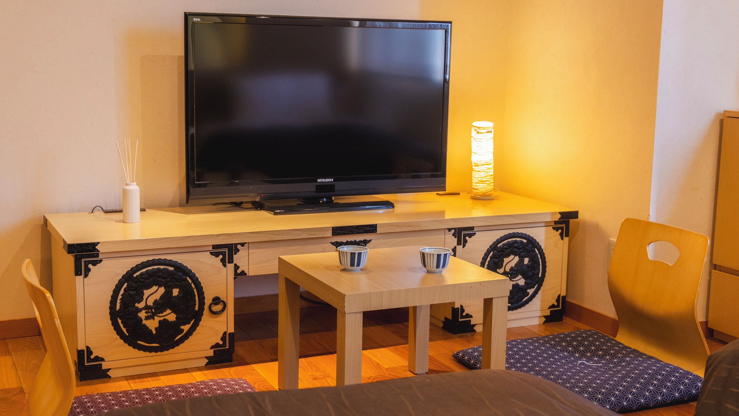 Quarto com televisão sobre um armário de madeira, uma mesinha com duas chávenas, uma cadeira e um candeeiro decorativo.