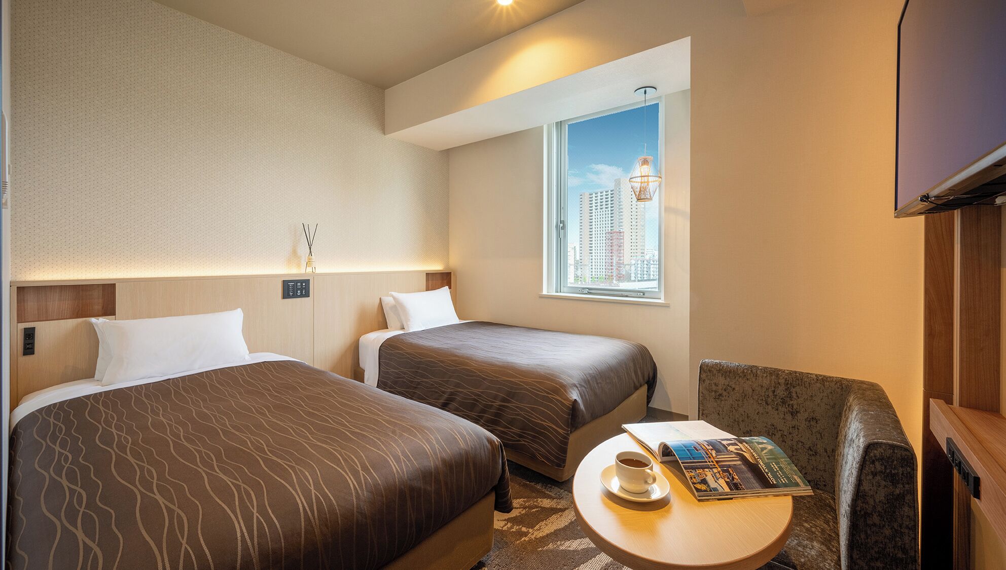 Hotelkamer met twee bedden, een klein tafeltje met een tijdschrift, een stoel, een raam met uitzicht op de stad en een bedieningspaneel aan de muur.