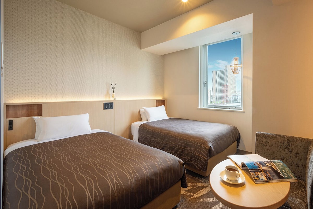 A hotel room with two beds, a small table with a magazine, a chair, a window with a city view, and a wall-mounted control panel.
