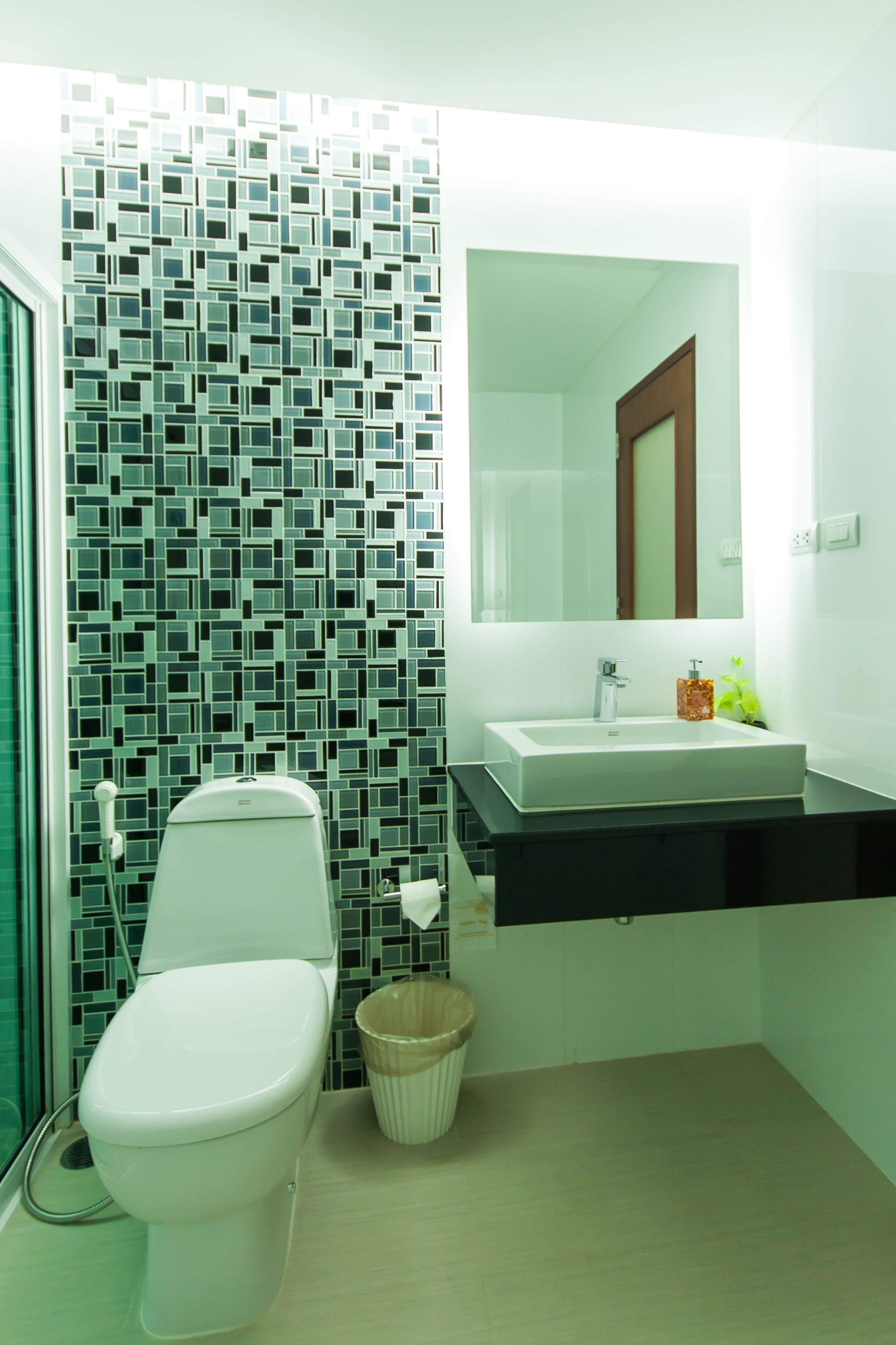 A modern bathroom with a tiled wall, a white toilet, a sink with a mirror, and a small trash bin.