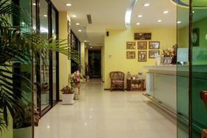 A hotel reception area with a reception desk, chairs, potted plants, and framed pictures on the wall.