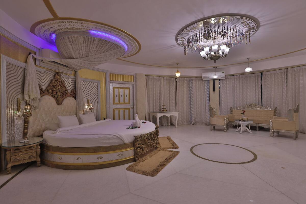 A luxurious bedroom with a large bed, ornate ceiling, chandelier, and elegant furniture.