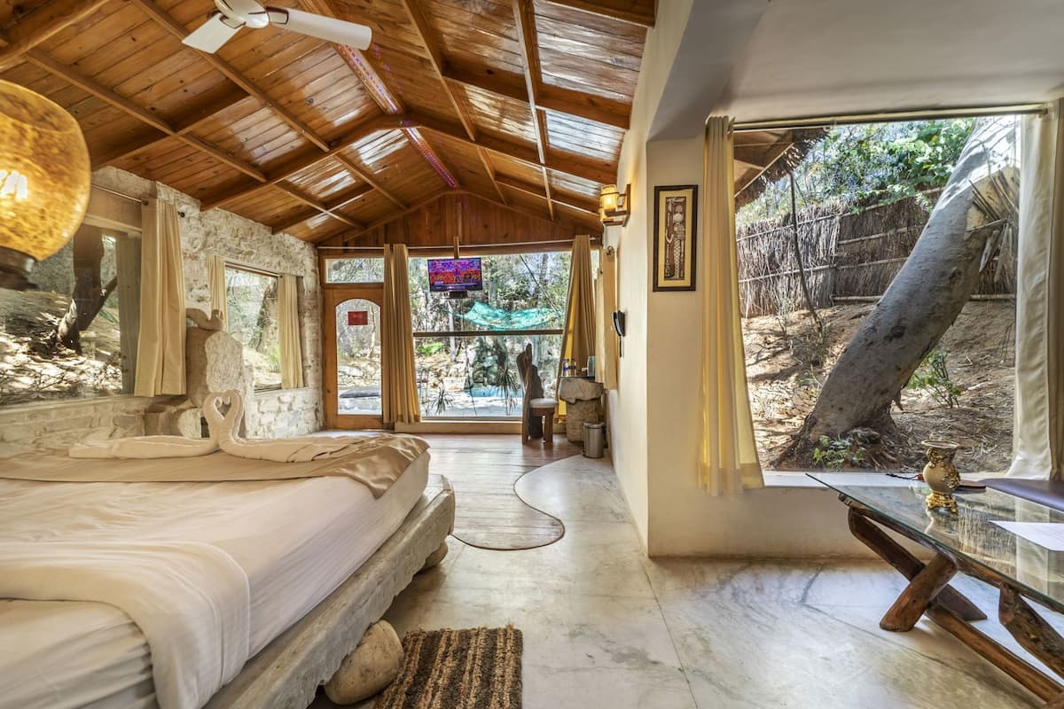 A room with a bed, a stone wall, a wooden ceiling, and a view of the outdoors.
