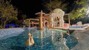 A nighttime view of a pool area with a fountain, surrounded by trees and a building with illuminated blue lights. - Lohagarh Fort Resort (Amer)