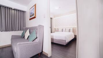 A hotel room with a grey sofa, a bed with white bedding, and a large window with grey curtains.