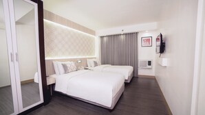 A hotel room with two beds, a large mirror, a wardrobe, a bedside table, a wall-mounted TV, and a window with curtains. - The Charter House (Makati)