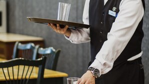A waiter holding a tray with a glass and a bottle, standing at a table with a cup and salt shakers. - The Charter House (Makati)