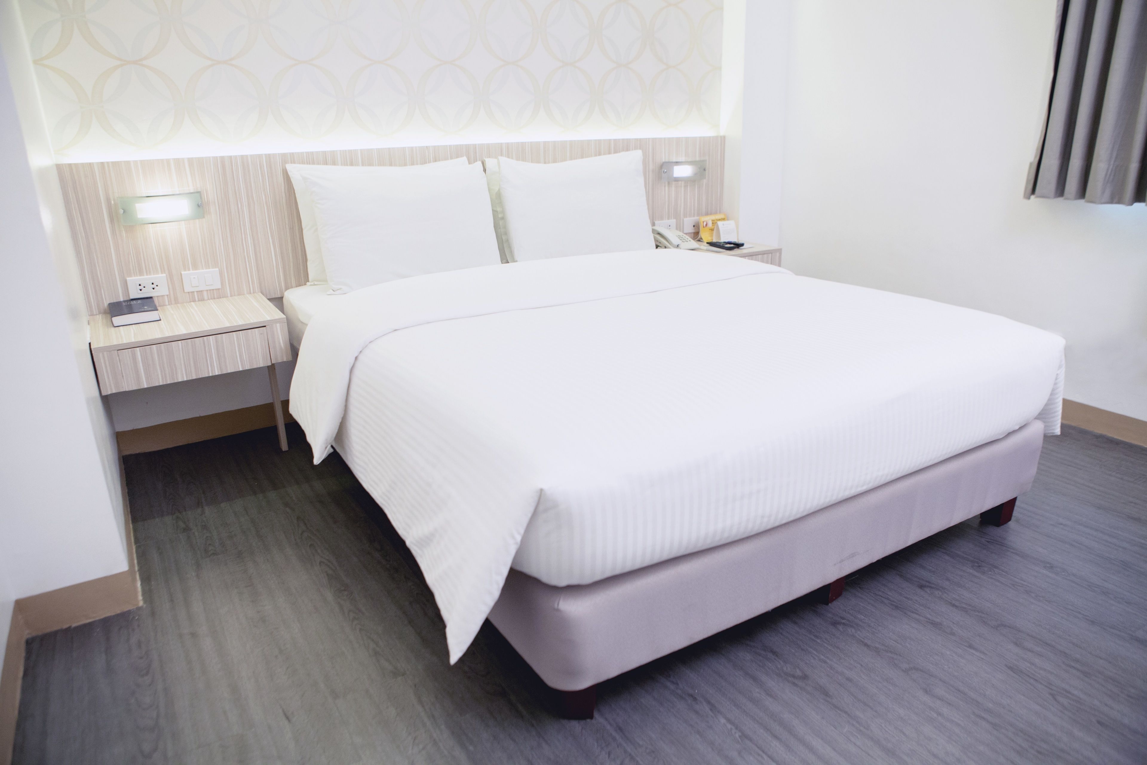 A neatly made bed with white linens, a bedside table with a lamp, and a wall-mounted light fixture.