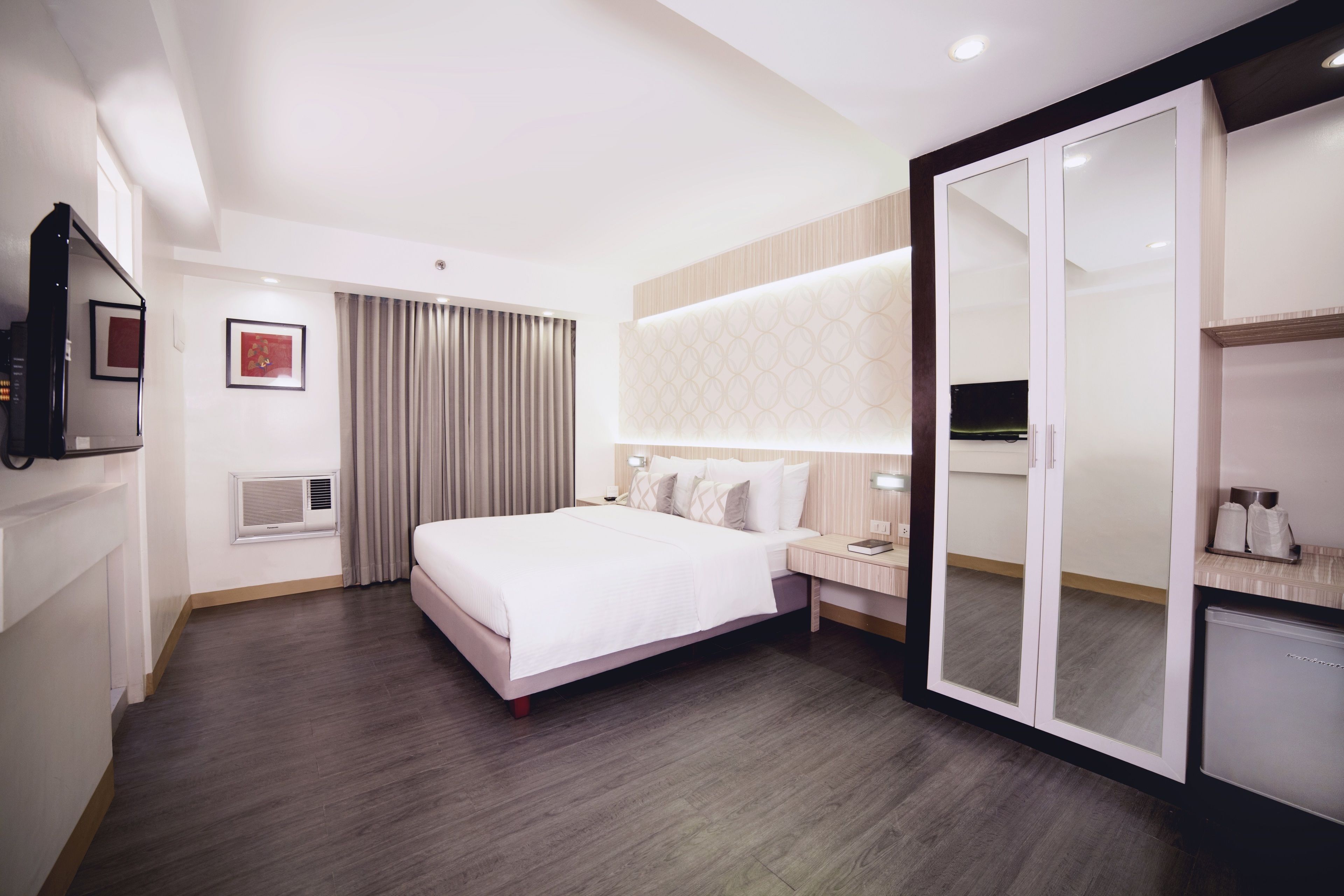 Habitación de hotel moderna con una cama grande, un armario con espejo, una mesita de noche con lámpara y un televisor de pared.