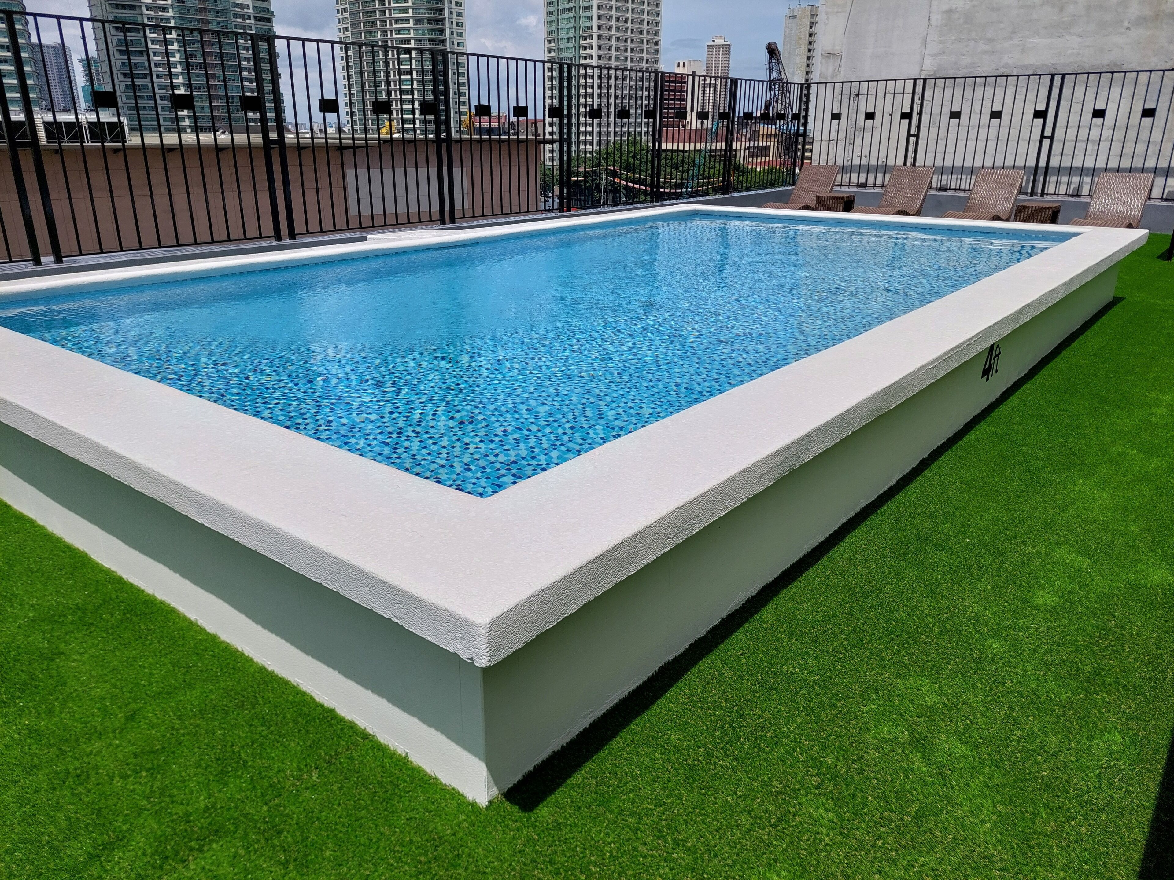A rectangular swimming pool with a white edge, surrounded by artificial green grass and a black metal fence.