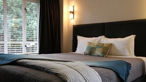Una cama bien tendida con un cabecero oscuro, una ventana con persianas y una lámpara de pared.
