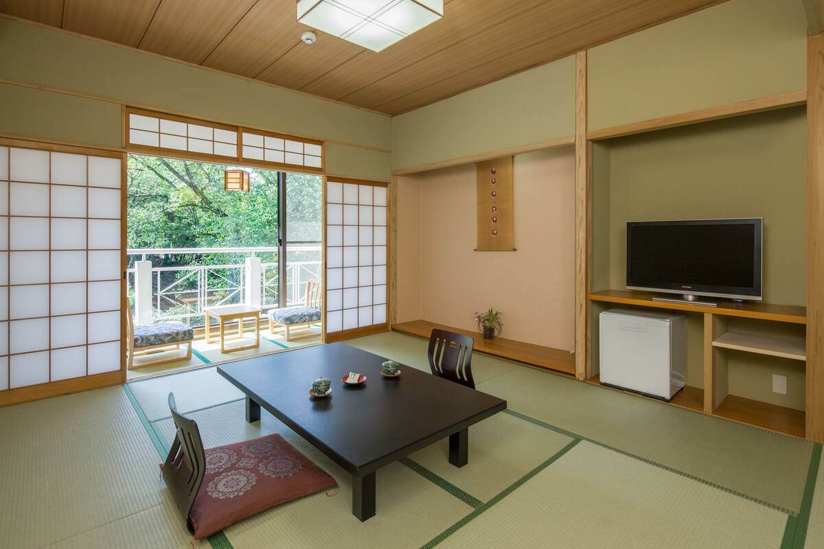 Ein traditioneller japanischer Raum mit Tatami-Böden, einem niedrigen Tisch mit Kissen, einem Fernseher und einem kleinen Kühlschrank.
