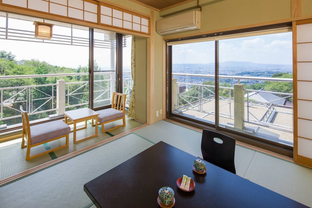 Ein traditioneller japanischer Raum mit Tatami-Böden, einem niedrigen Tisch mit Kissen und Blick auf eine Stadtlandschaft durch große Fenster.