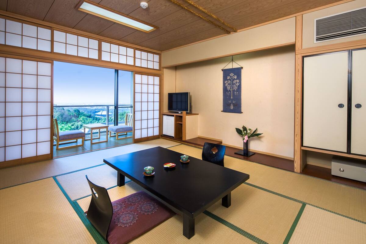 Ein traditioneller japanischer Raum mit Tatami-Böden, einem niedrigen Tisch mit Kissen, einem Fernseher und einer Schiebetür zu einem Balkon mit Blick auf Grünflächen.