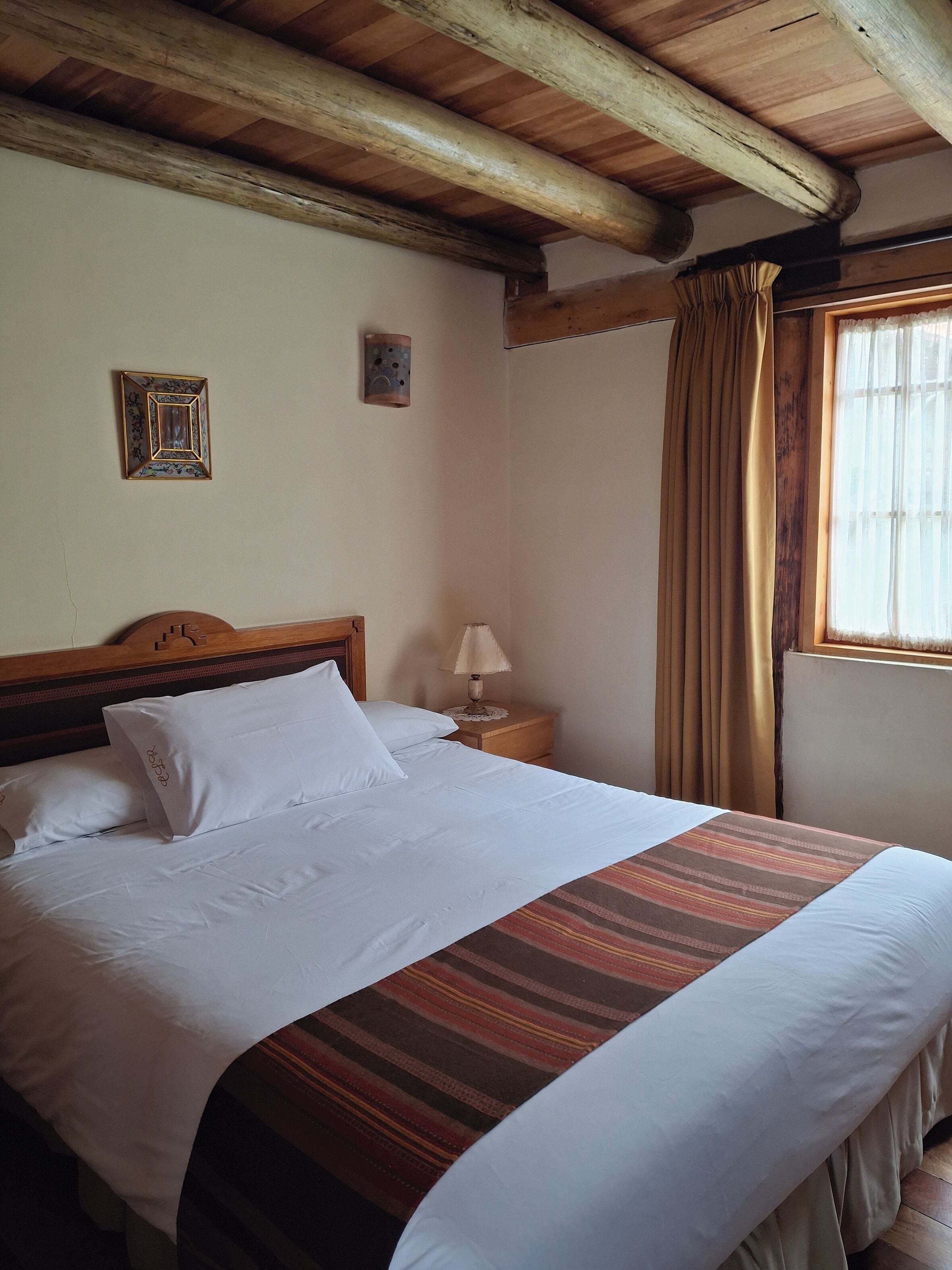Cama bem arrumada com lençóis brancos e manta listrada, cabeceira de madeira, mesa de cabeceira com abajur e janela com cortinas.