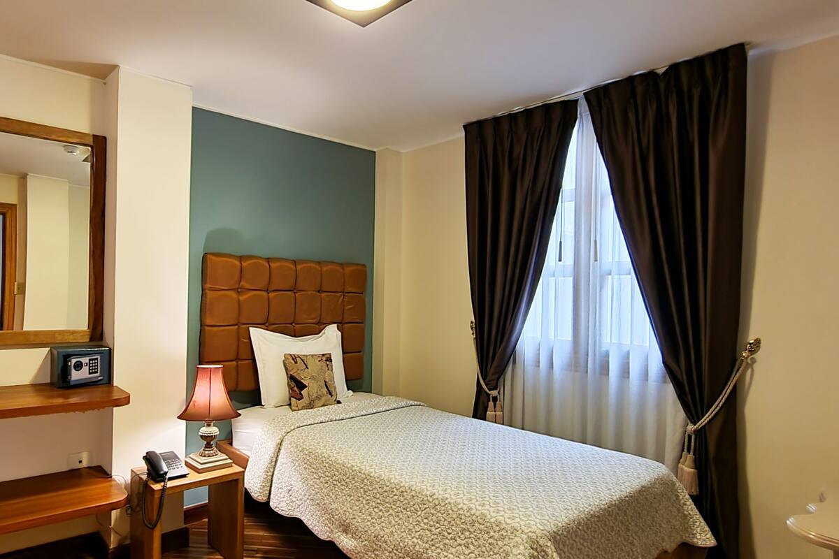 A hotel room with a bed, a bedside table with a lamp, a telephone, a mirror, and a window with curtains.