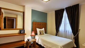 Habitación de hotel con cama, mesita de noche con lámpara, teléfono, espejo y ventana con cortinas.
