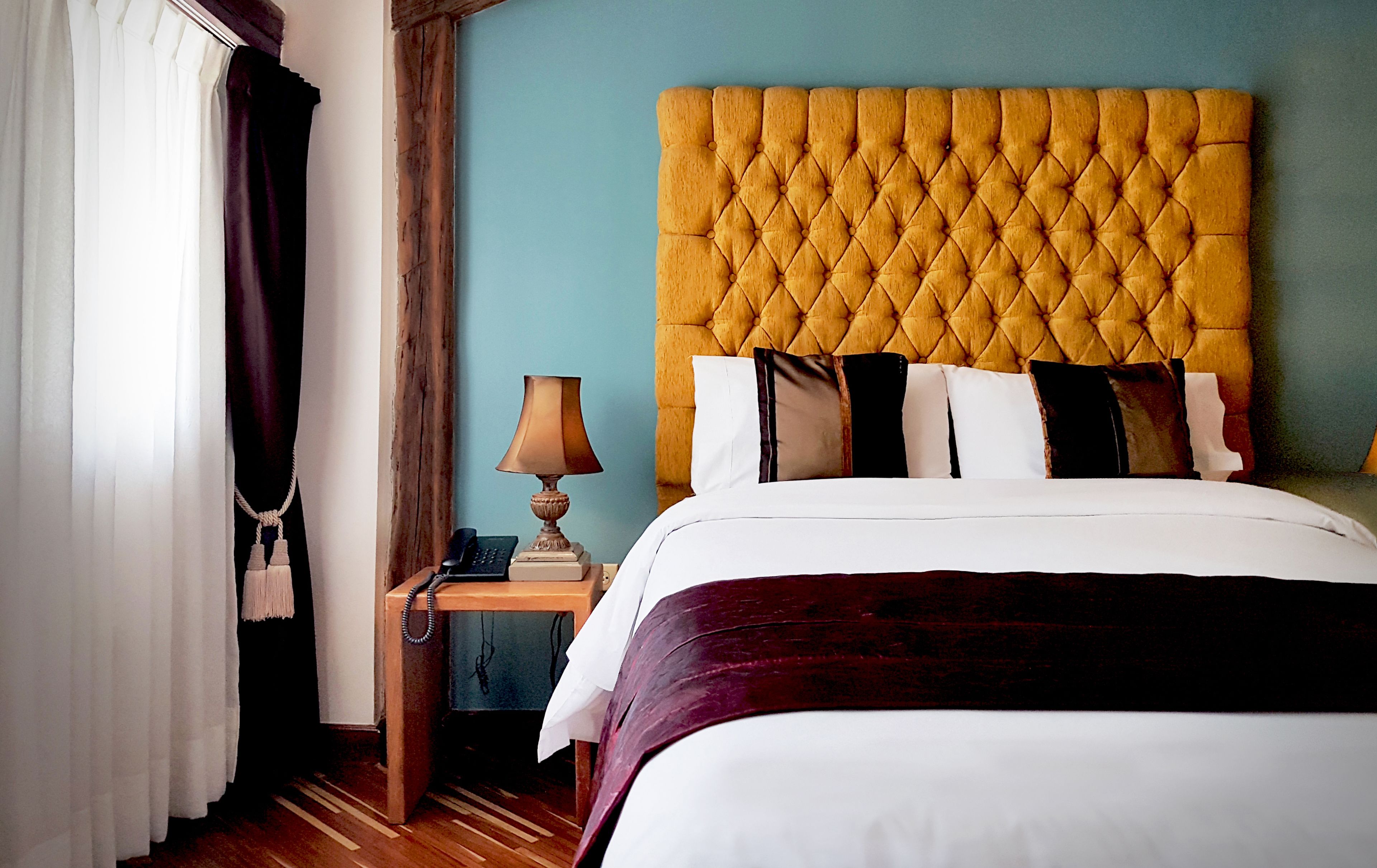 A hotel room with a tufted headboard, a bedside table with a lamp, and a bed with white bedding and multiple pillows.